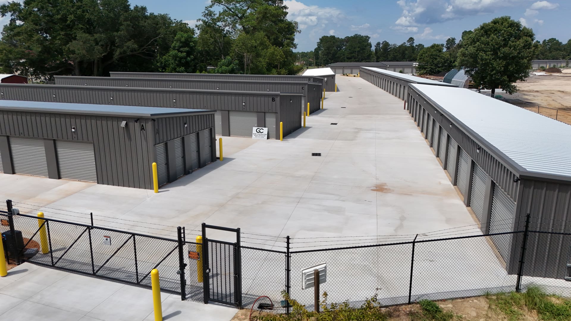 A wide concrete drive aisle at My Storage running between two rows of grey roll-up unit doors, with the site's black perimeter fence and gate in the foreground.