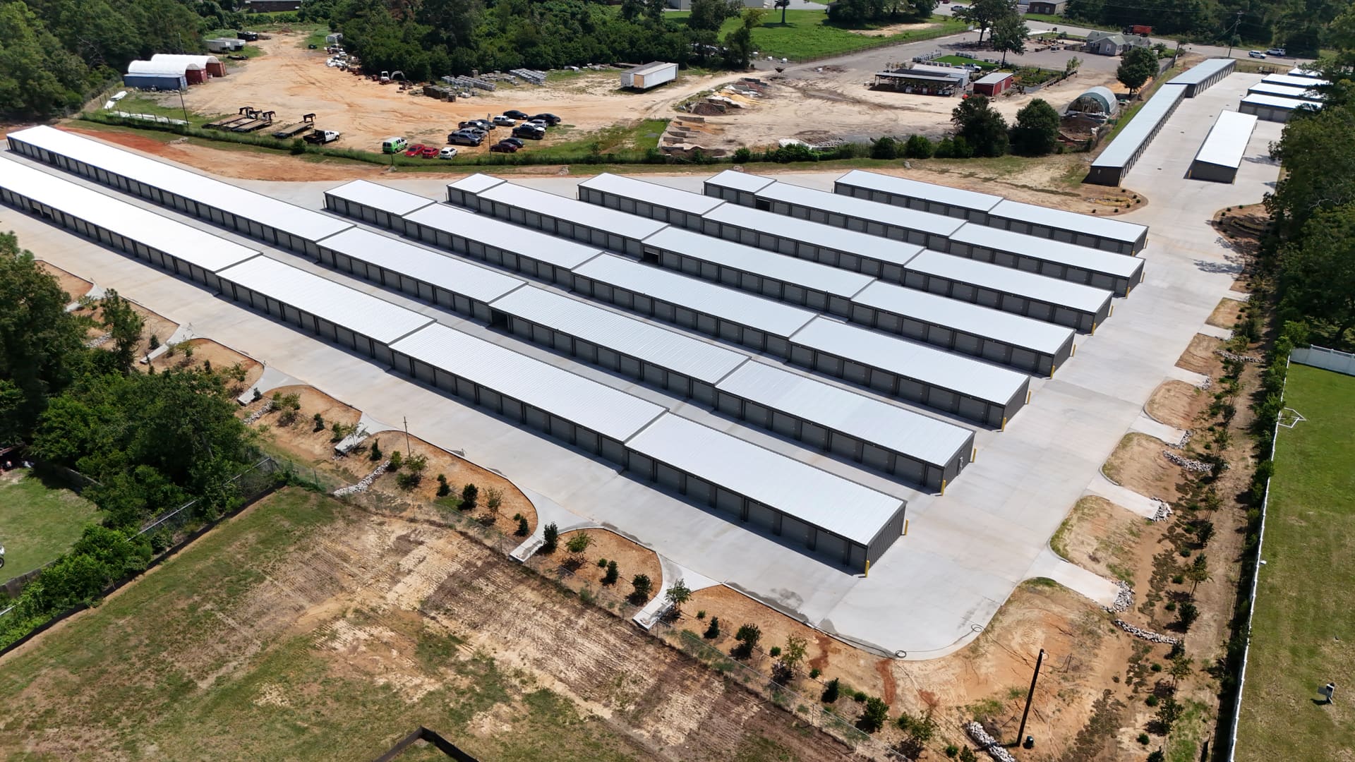 Aerial view across My Storage: rows of drive-up storage buildings with white roofs and grey roll-up doors, separated by wide concrete drive aisles, with the fenced boundary and trees around the site.